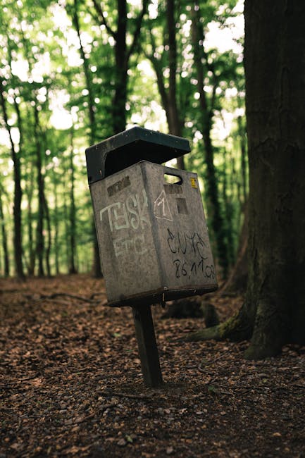 A weathered grey metal mailbox with graffiti tags on its surface, mounted on a tilted, black metal post. The mailbox is situated in a wooded area with tall trees and green foliage in the background, and the ground covered in brown soil and leaf litter. The mailbox's lid is open, revealing a dark interior, and it appears to be in a state of disrepair with visible graffiti and signs of exposure to the elements. The scene captures an outdoor environment that could relate to private property or alternative waste handling, with natural light filtering through the tree canopy. Rubbish Removal Stockwell often deals with clearing bulky waste from similar outdoor areas, including removing discarded boxes, old equipment, or other debris left in such settings.