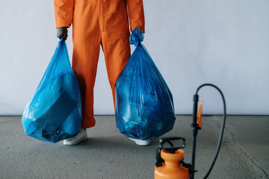 A person dressed in orange overalls and white shoes is standing on a concrete floor, holding two large blue plastic garbage bags filled with waste, one in each hand. The bags are semi-transparent, revealing various types of rubbish inside, and are tightly knotted at the tops. The background features a plain, light-colored wall, with part of a black and orange handheld sprayer or pressure spray device visible in the foreground. The scene suggests an active waste collection or rubbish removal process, typical of independent waste handling services such as those provided by Rubbish Removal Stockwell, aimed at clearing bulky waste items from residential or commercial premises on-site.