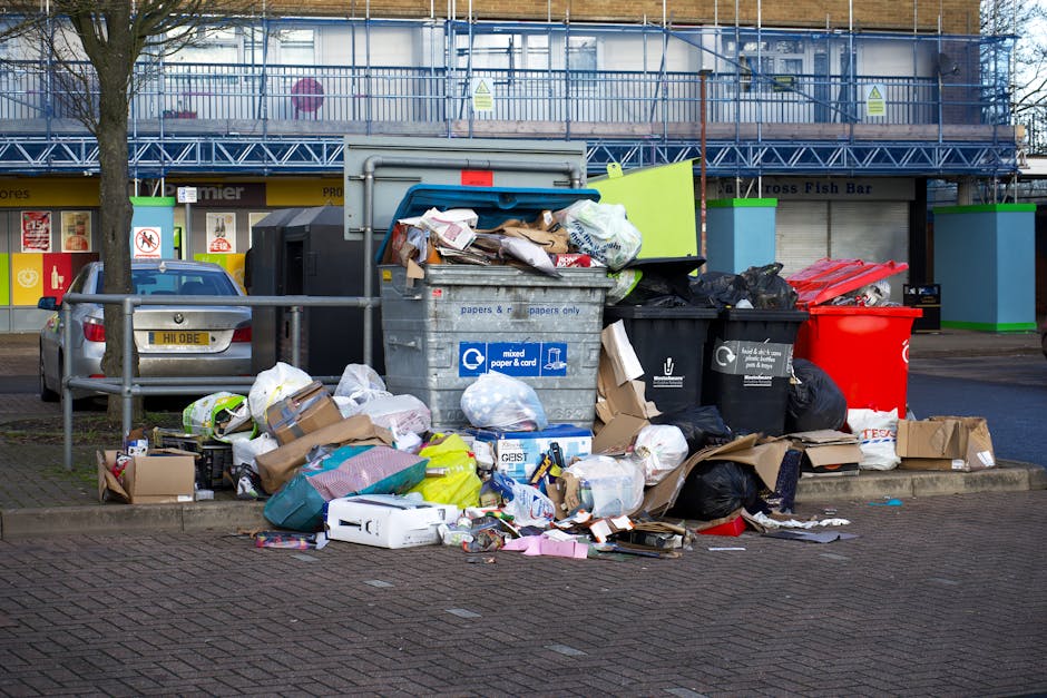 An outdoor scene showing a large accumulation of mixed waste and rubbish overflowing from multiple wheelie bins and bags placed on a paved sidewalk area. The waste includes crumpled cardboard boxes, plastic packaging, paper bags, and miscellaneous items such as a white plastic container and some textiles. The bins include a grey mixed paper and cardboard bin, a black general waste bin, and a red recycling bin, all positioned near a waist-high metal railing. In the background, a row of shops with partially visible signs, windows, and corporate branding is visible, along with parked cars on the left side. Behind the shopping units, there is a building under construction or renovation, surrounded by scaffolding and protective netting. The scene is illuminated by natural daylight, emphasizing the cluttered state of the rubbish, which appears to be a result of an uncontrolled or unbooked waste collection, highlighting the importance of private rubbish removal services such as those offered by Rubbish Removal Stockwell.