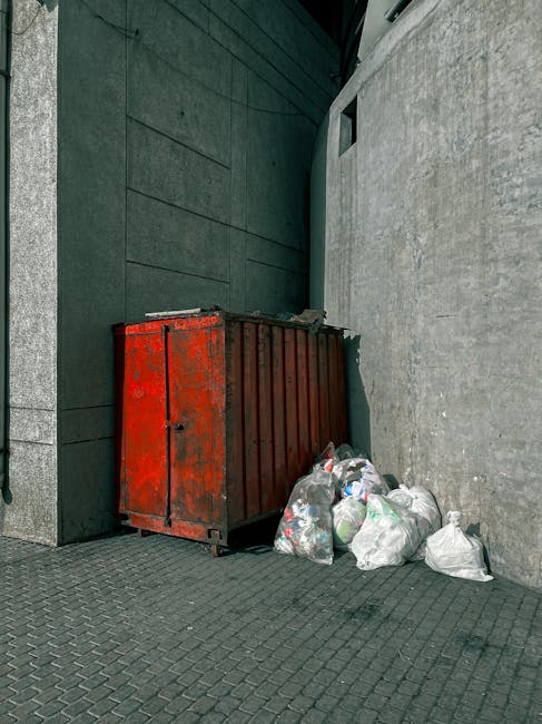 A red, weathered metal dumpster with a ribbed surface design is positioned against a dark green wall and a light grey concrete wall on a paved sidewalk. The dumpster appears to be used for waste storage and disposal, with its paint showing signs of rust and wear. Several tightly tied white plastic garbage bags filled with household or office waste are stacked next to the dumpster, leaning slightly against it. The garbage bags seem full and are likely prepared for collection or an alternative private waste disposal service. The scene is outdoors, with ambient natural light illuminating the area, and the setting suggests an urban environment, possibly an alleyway or side street. This visual composition aligns with the activities of rubbish removal services, including both on-site clearance and independent rubbish collection, as provided by companies such as Rubbish Removal Stockwell, operating in areas like South Lambeth Road. The surroundings indicate a practical waste management point, typical for local waste handling outside commercial or residential buildings.