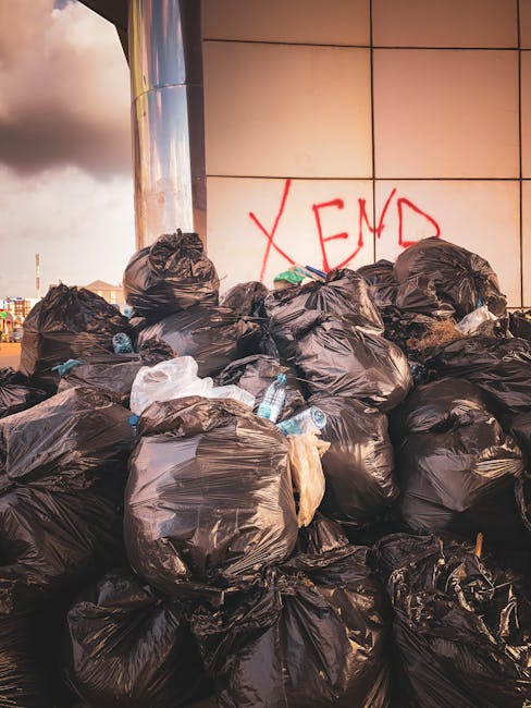 A large collection of black plastic rubbish bags piled against a modern building with reflective, beige-colored panels. The bags are filled with waste and some are torn or bulging, revealing plastic bottles, paper, and other refuse inside. Among the bags, several clear plastic bottles are visible, some partially crushed. The bags are arranged haphazardly on a paved surface, likely on a street or alleyway, with environmental elements like cloudy sky and distant urban structures in the background. The wall behind the rubbish has graffiti in red spray paint that reads 'XEND', adding an urban context to the scene. This scene demonstrates an informal, unmanaged waste accumulation typical of situations where private rubbish removal services such as those provided by Rubbish Removal Stockwell are needed for on-site rubbish clearance and alternative waste handling. The overall environment hints at a business or residential area where waste disposal is temporarily delayed or awaiting collection, emphasizing the importance of prompt rubbish removal and waste management solutions.