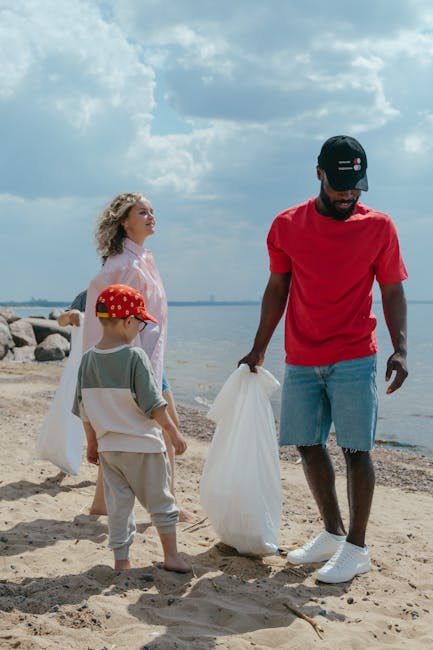 A group of three individuals with light skin, consisting of a young woman, a young boy, and a man, are on a sandy beach near a body of water under a partly cloudy sky. The woman has curly blonde hair and is wearing a light pink or white blouse, standing slightly behind the others. The young boy, dressed in light-colored shorts and a grey t-shirt, is wearing a red cap with a pattern and is carrying a white rubbish bag. The man, wearing a red t-shirt, light blue denim shorts, white sneakers, and a black cap with a small red and white logo, is holding a white rubbish bag and appears to be preparing to collect waste or litter. Behind them, the shoreline is lined with rocks and the water extends toward the horizon. The scene suggests a community or family effort in cleaning up the environment, with the visible trash bags hinting at a process of waste collection or disposal, possibly coordinated by a rubbish removal service such as Rubbish Removal Stockwell, highlighting an outdoor private disposal or clean-up activity.